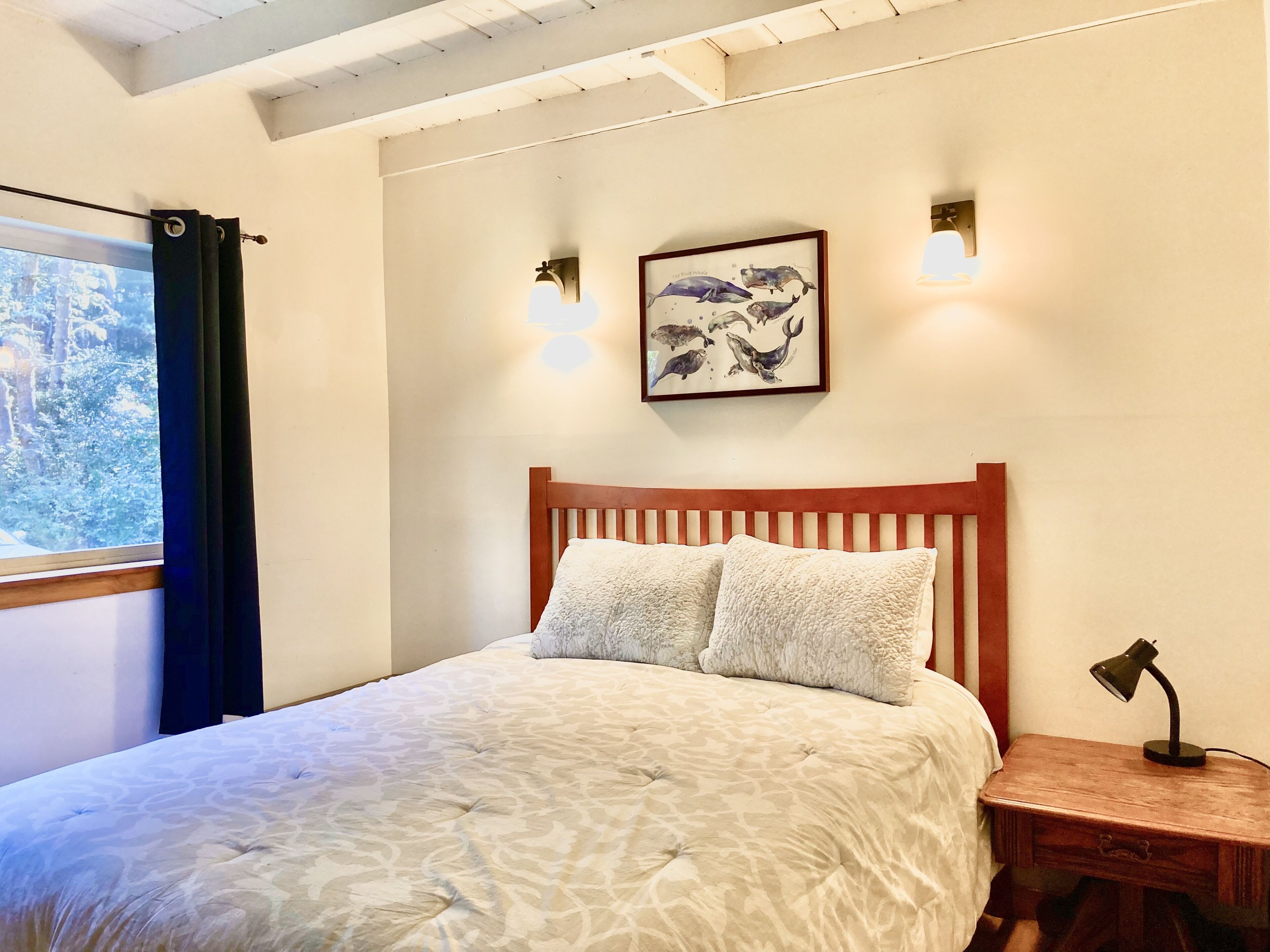 A bedroom with a bed, a nightstand, a wall-mounted picture of whales, and a window with curtains.