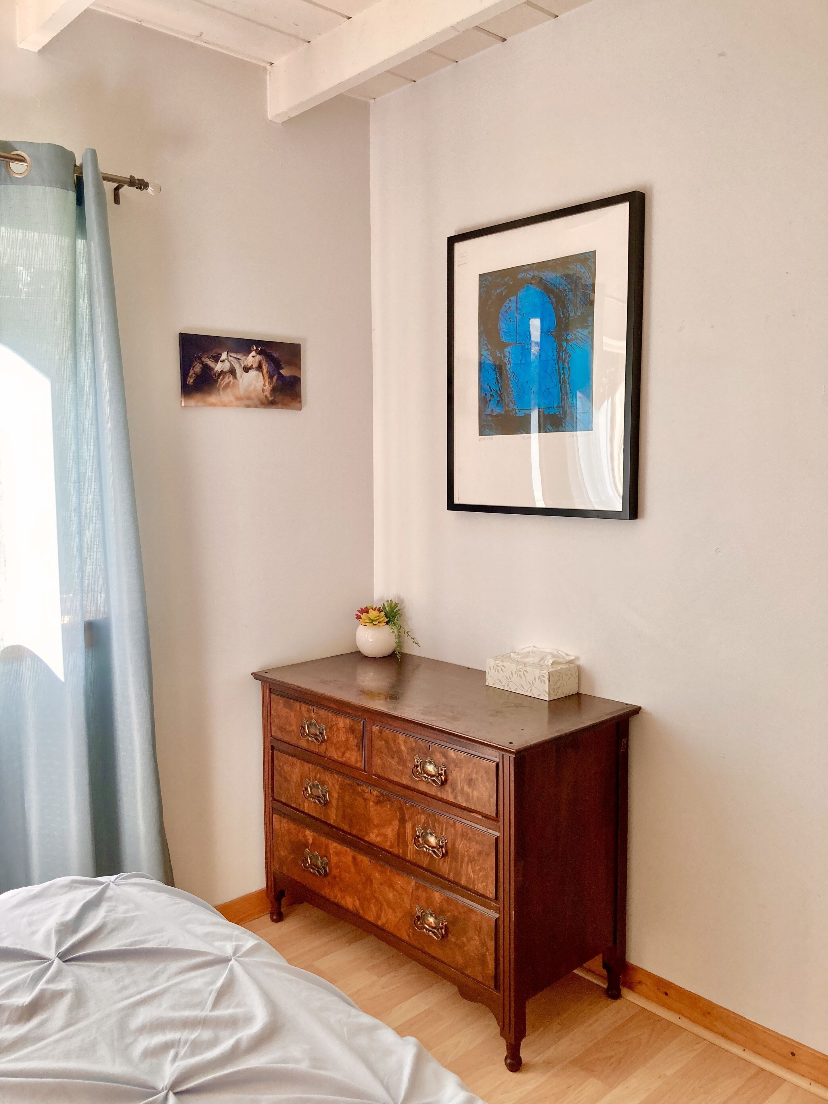 A bedroom with a wooden dresser, a framed abstract painting, a small vase with flowers, and a bed with a white cover.
