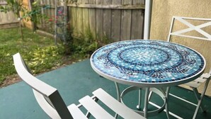 Un tavolo esterno rotondo con un piano in mosaico, circondato da sedie bianche, posizionato in un patio verde con una recinzione in legno sullo sfondo.
