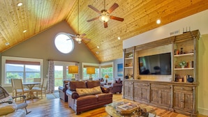 A spacious living room with a high ceiling, wooden beams, a large TV, leather sofas, and a glass coffee table. - Luxury Home, Stunning Views!, One-Level, Near Main St., Privacy, FirePit & More (Waynesville)