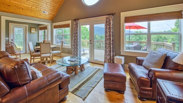 A spacious living room with leather sofas, a glass coffee table, and a view of a deck. - Luxury Home, Stunning Views!, One-Level, Near Main St., Privacy, FirePit & More (Waynesville)