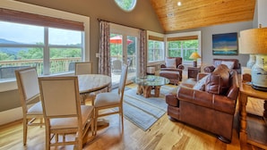 A cozy living room with a wooden ceiling, a round table, leather sofas, and large windows offering a view of the outdoors. - Luxury Home, Stunning Views!, One-Level, Near Main St., Privacy, FirePit & More (Waynesville)