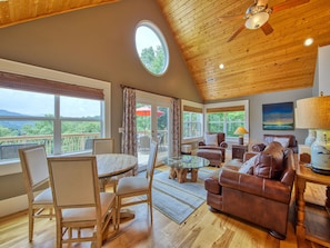 A cozy living room with a wooden ceiling, a round table, leather sofas, and large windows offering a view of the outdoors.