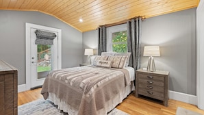 A bedroom with a wooden ceiling, a bed with a brown blanket, two bedside lamps, a nightstand, and a window with curtains. - Luxury Home, Stunning Views!, One-Level, Near Main St., Privacy, FirePit & More (Waynesville)
