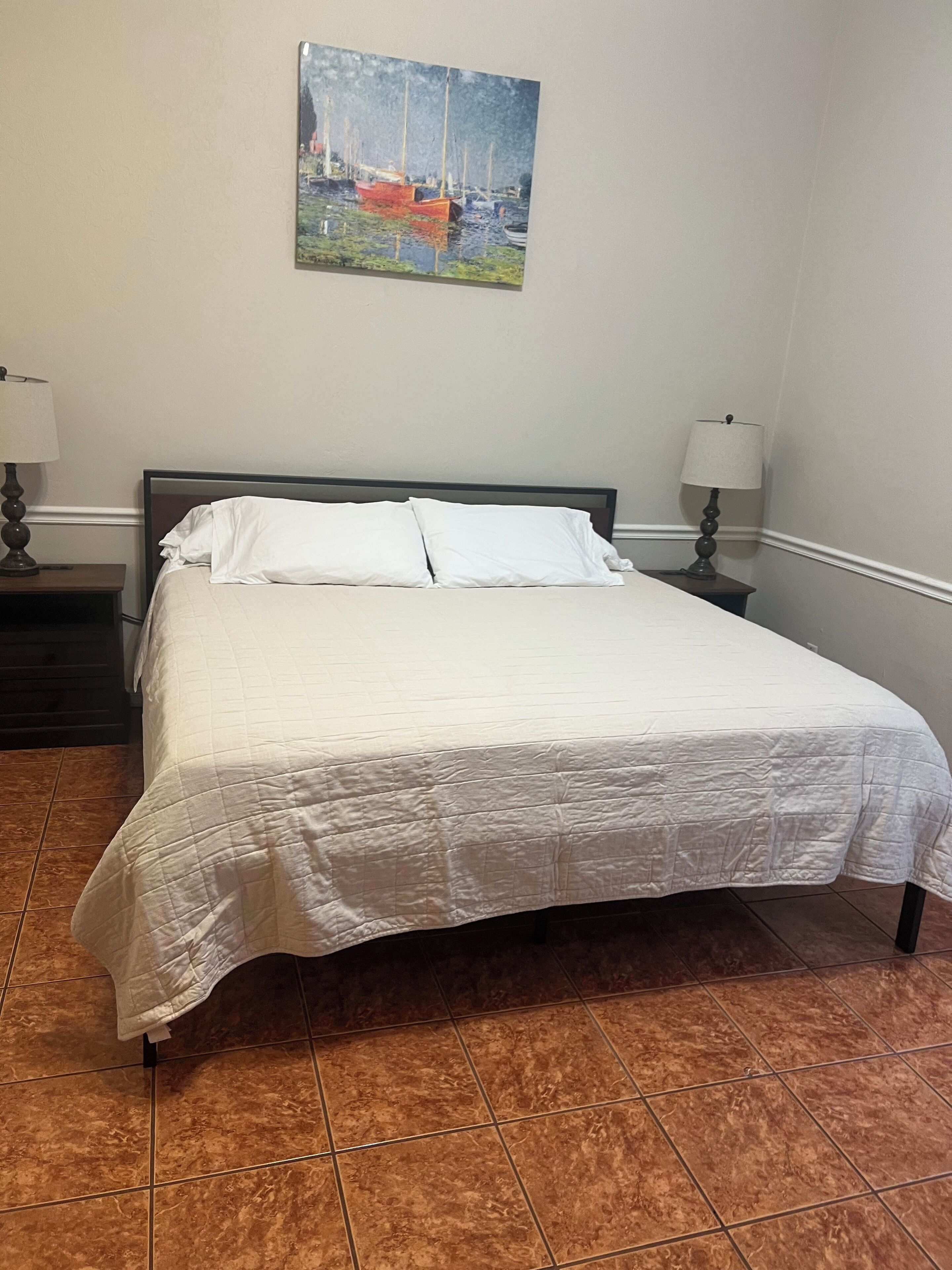 Een netjes opgemaakt bed in een kamer met een schilderij van boten aan de muur, een nachtkastje met een lamp en een betegelde vloer.