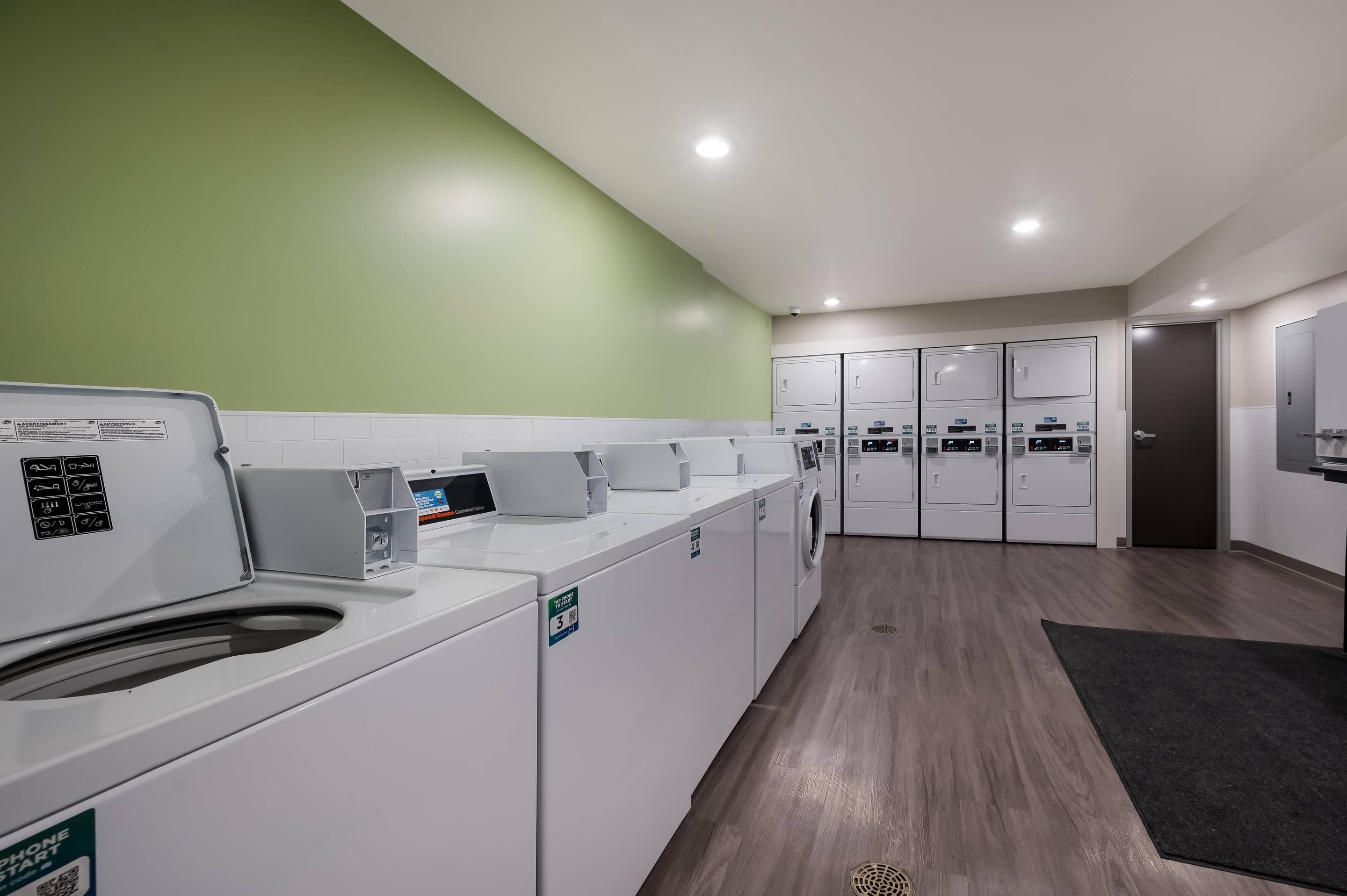 A laundry facility with multiple washing machines and dryers, a green wall, and a wooden floor.