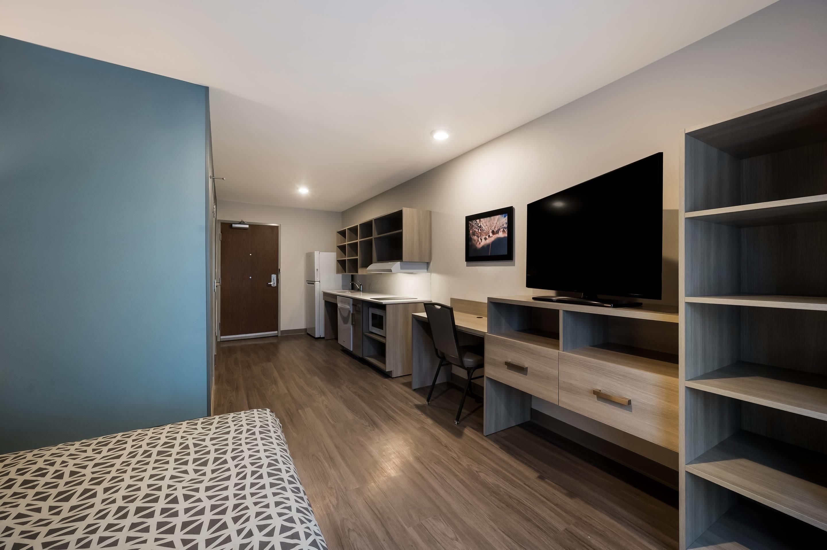 Un espacio habitable moderno y compacto con kitchenette, comedor y un televisor grande montado en la pared.