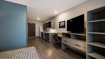 Un espacio habitable moderno y compacto con kitchenette, comedor y un televisor grande montado en la pared.