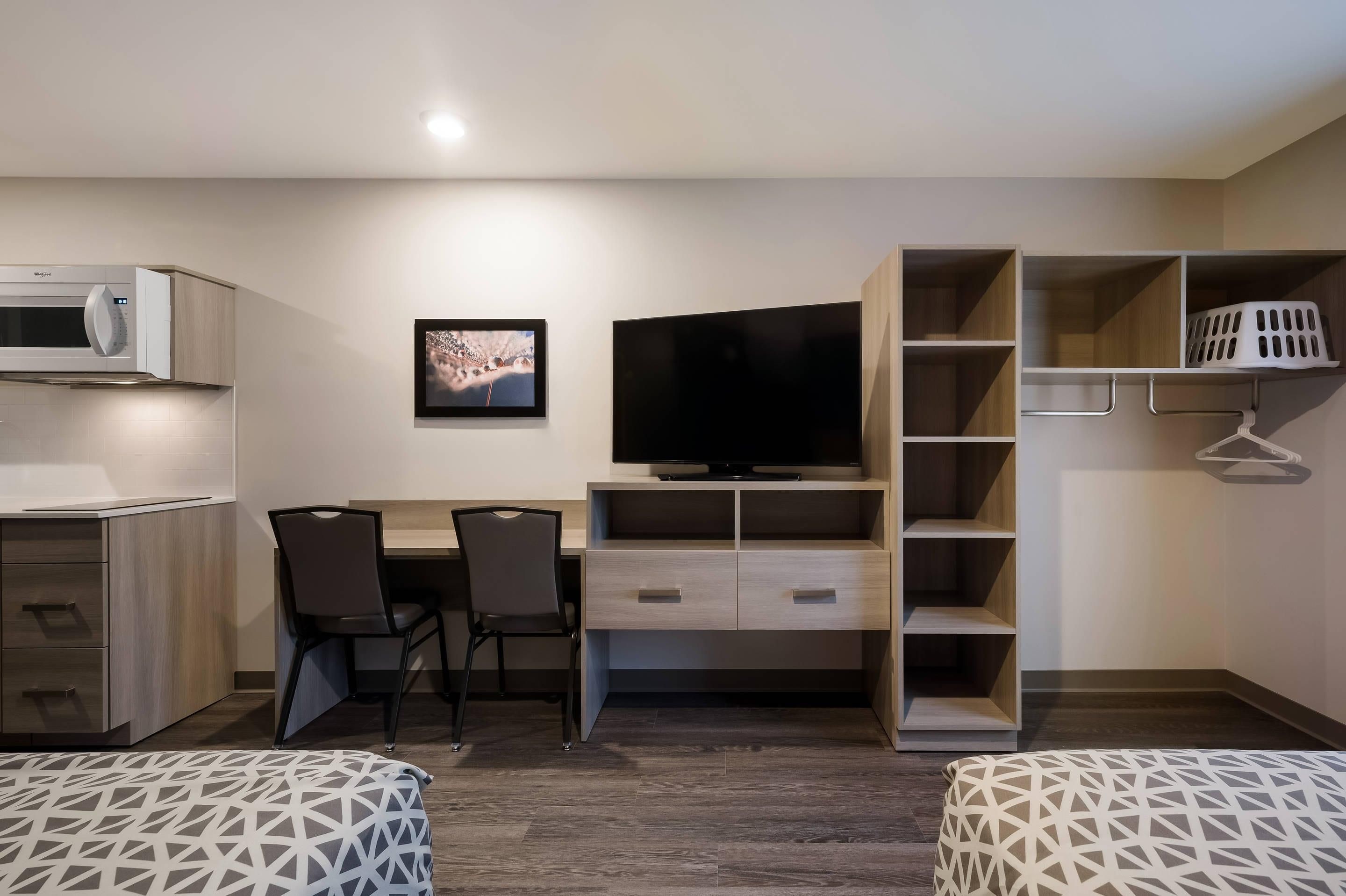 A modern hotel room with a kitchenette, a flat-screen TV, a desk with two chairs, a wardrobe, and a built-in shelving unit.