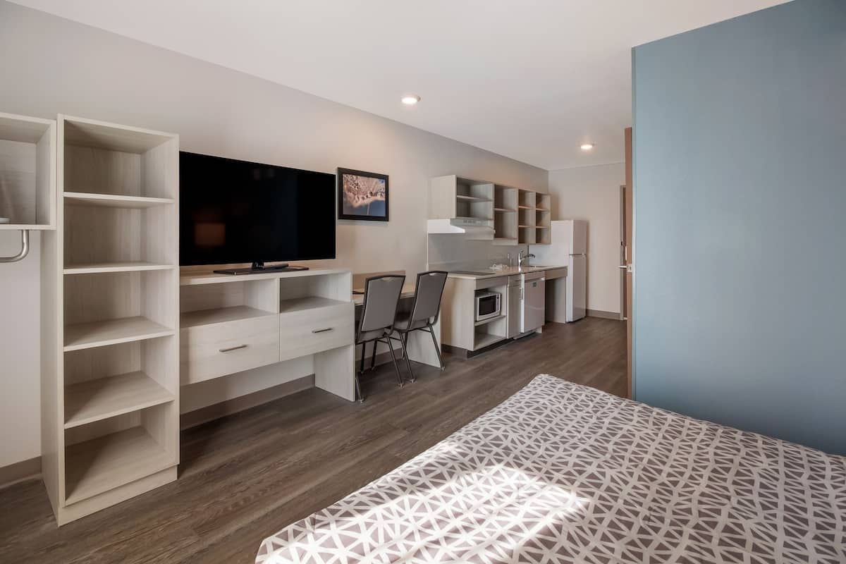 A modern bedroom with a bed, a TV, a desk, and a kitchenette.