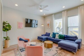 Ein modernes Wohnzimmer mit einem blauen Ecksofa, einem rosa Sessel, einem runden Couchtisch und einem Deckenventilator.