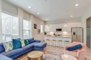 Uma sala de estar moderna com um sofá seccional azul, uma poltrona rosa, uma mesa de centro redonda e uma área de cozinha com armários brancos e uma geladeira de aço inoxidável.