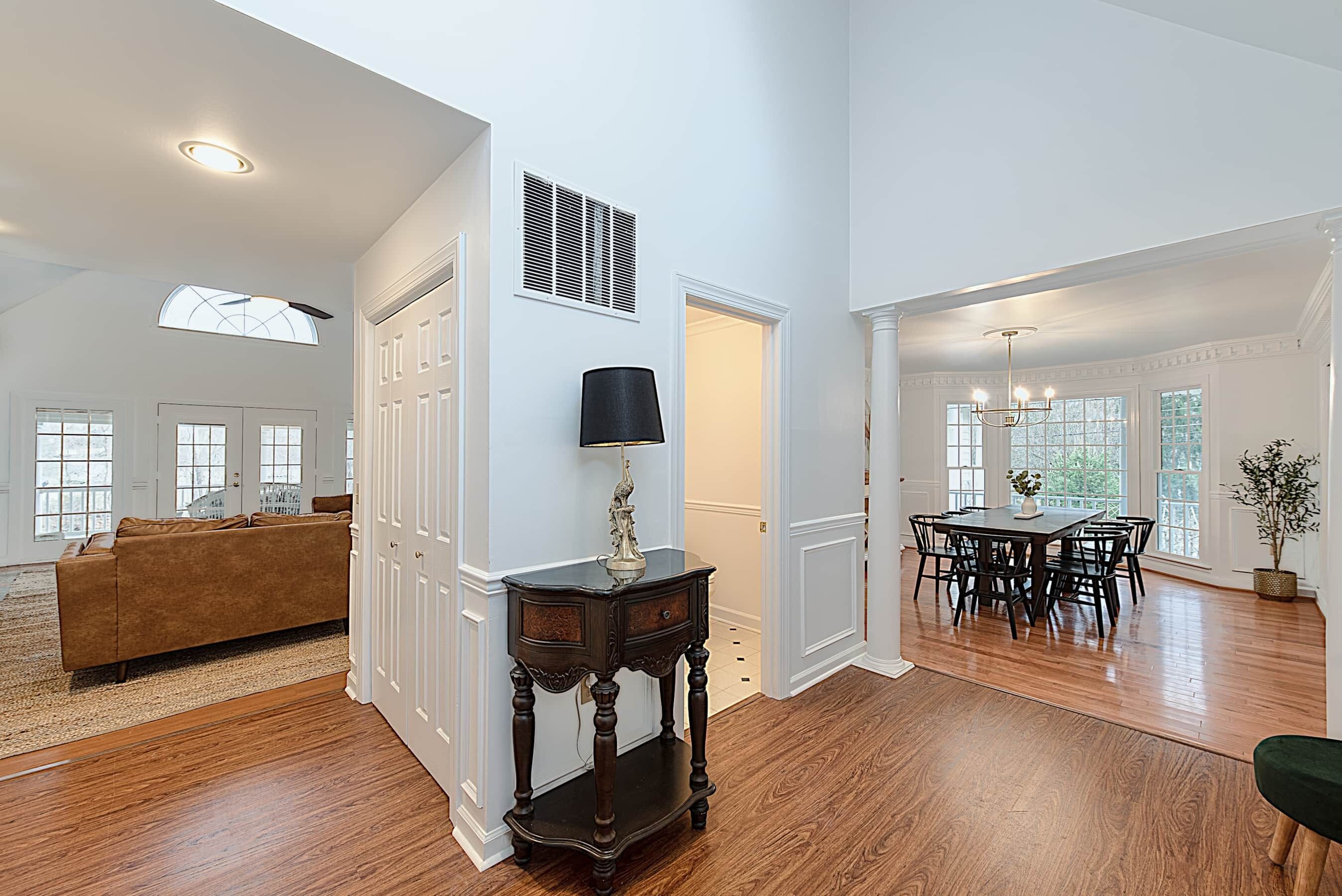A spacious living room with hardwood floors, a brown sofa, a dark wooden side table with a lamp, and a dining area with a table and chairs.