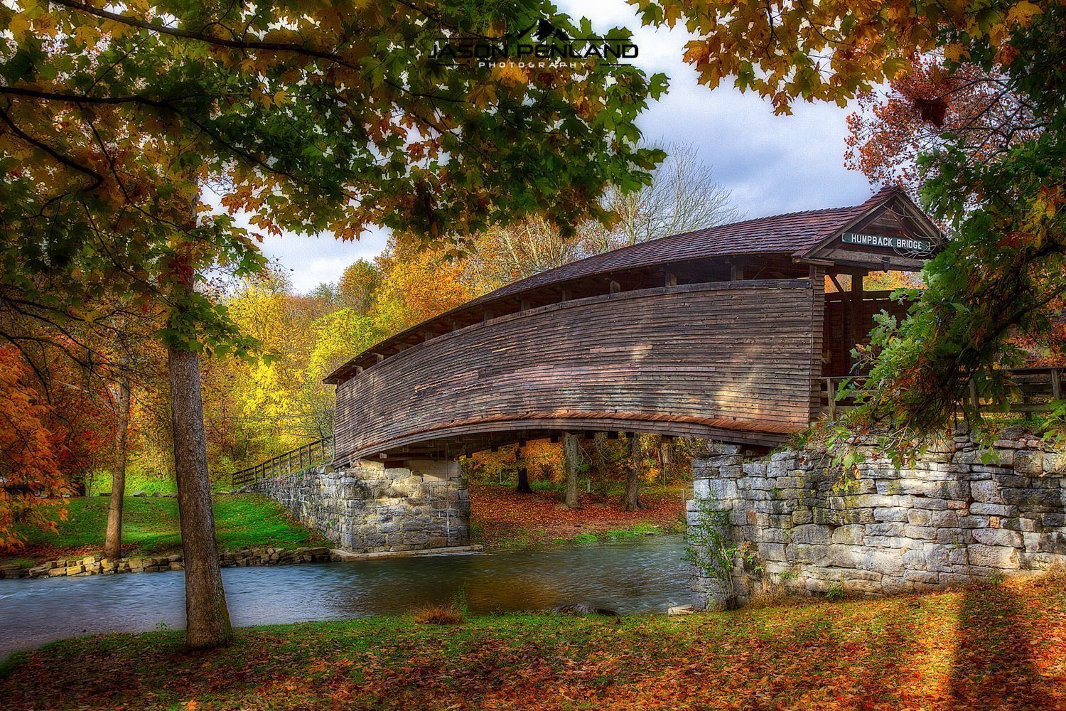 A wooden covered bridge over a river, surrounded by trees with autumn foliage.