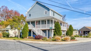 A two-story house with a front porch, a small staircase, and a garage. - Beach Haven | Walk to Downtown Chincoteague, Wraparound Deck, Smart TVs & Garage for Beach Gear (Chincoteague Island)