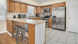 A kitchen with wooden cabinets, a stainless steel refrigerator, a microwave, a stove, and a sink. There are two bar stools and a window with a curtain. - Beach Haven | Walk to Downtown Chincoteague, Wraparound Deck, Smart TVs & Garage for Beach Gear (Chincoteague Island)