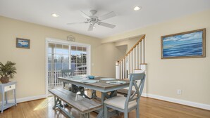 A dining area with a wooden table and benches, a ceiling fan, and a view of the outdoors through a glass door. - Beach Haven | Walk to Downtown Chincoteague, Wraparound Deck, Smart TVs & Garage for Beach Gear (Chincoteague Island)