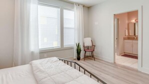 Ein Schlafzimmer mit Bett, einem Sessel, einer Topfpflanze und einem durch eine geöffnete Tür einsehbaren Badezimmer.