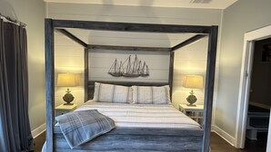 A four-poster bed with a nautical-themed headboard, two bedside tables with lamps, and a view of a hallway.
