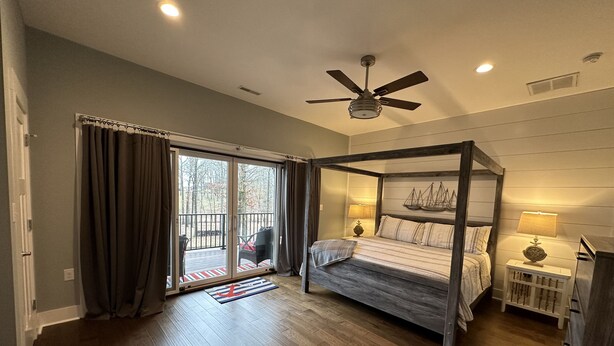 A bedroom with a four-poster bed, a ceiling fan, a nightstand with a lamp, and a view through a sliding glass door to an outdoor deck.