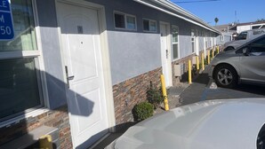 A row of single-story buildings with white doors and windows, a brick base, and a parked car. - La Perla Motel (South Gate)