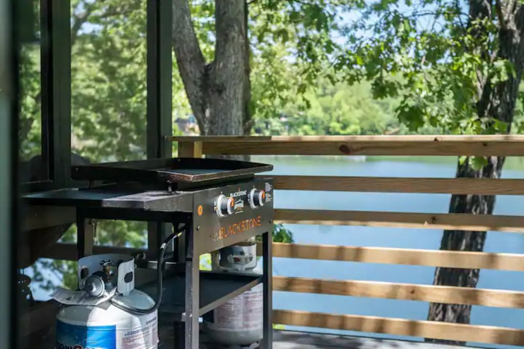 Un barbecue équipé de bouteilles de propane et doté d’une surface de cuisson plane, installé devant une terrasse en bois et un lac.