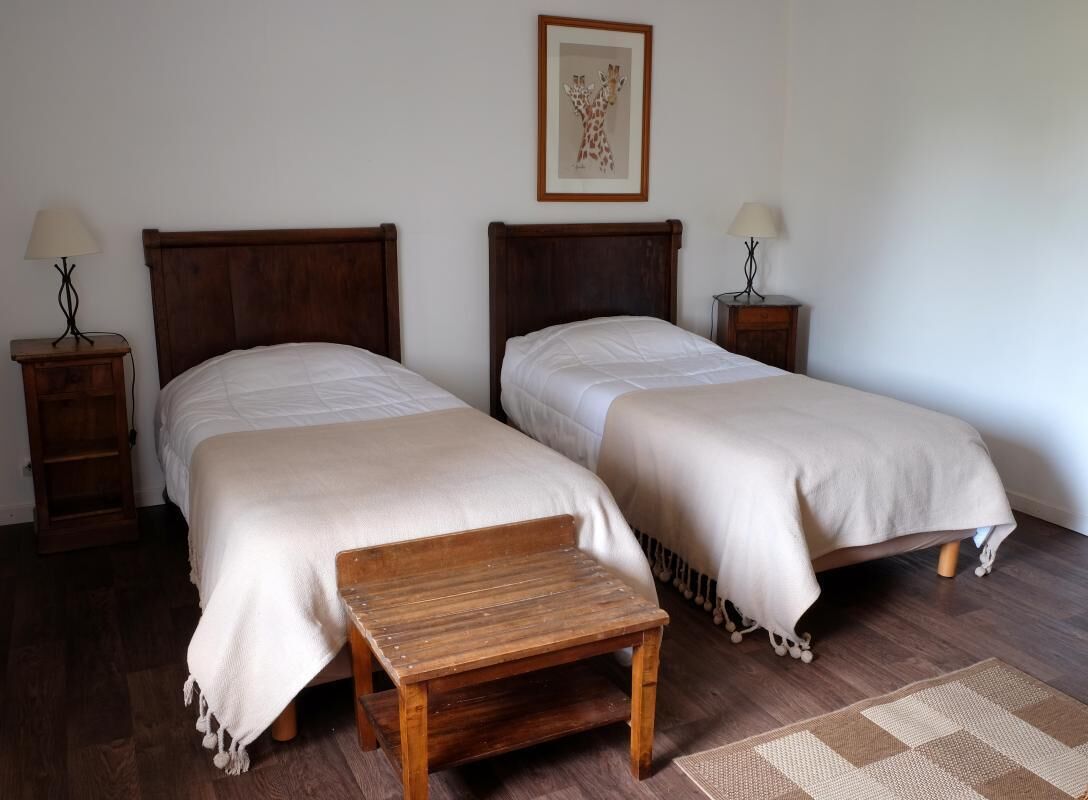 Ein Zimmer mit zwei Einzelbetten, Holz-Kopfteilen, Nachttischen inklusive Lampen, einer Holzbank und einem gewebten Teppich.