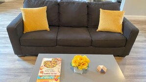 A dark brown sofa with two yellow cushions, a small table with a book, a vase of yellow flowers, and a geometric object. - High End Guest House by I-5, Bethel, Simpson (Redding)