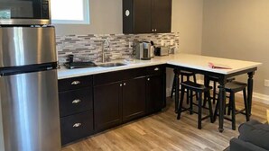 A modern kitchen with dark cabinetry, a stainless steel refrigerator, a tiled backsplash, and a wooden dining table with bar stools. - High End Guest House by I-5, Bethel, Simpson (Redding)