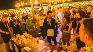 Um encontro social com pessoas ao redor de uma mesa com copos e garrafas, um bar ao fundo e uma placa que diz "OPEN BAR".