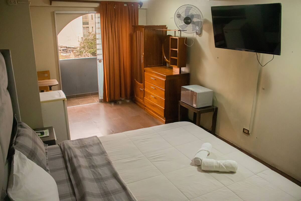 A hotel room with a bed, a wooden dresser, a TV mounted on the wall, and a fan.