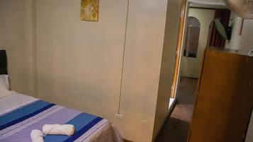 A hotel room with a bed, a wall-mounted power outlet, a door, and a fan.