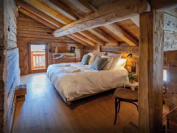 A cozy log cabin bedroom with a wooden bed, a nightstand, a chair, and a window with a view.