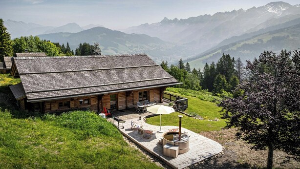 A rustic lodge with a wooden exterior, a deck, and a hot tub in the yard, surrounded by mountains. - Chalet Sisar - ski-in ski-out break for 12, La Clusaz - OVO Network (La Clusaz)