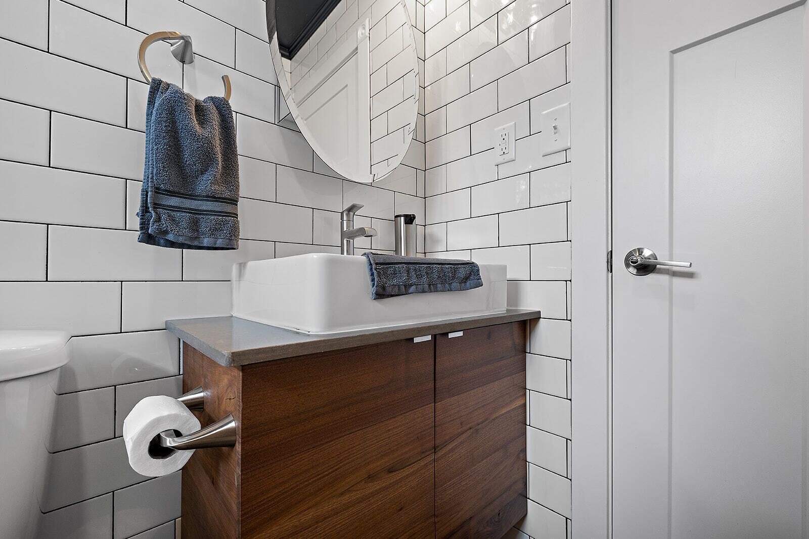 Une salle de bain avec des murs recouverts de carreaux blancs, un meuble-lavabo en bois, un lavabo blanc et une serviette suspendue à un porte-serviettes.