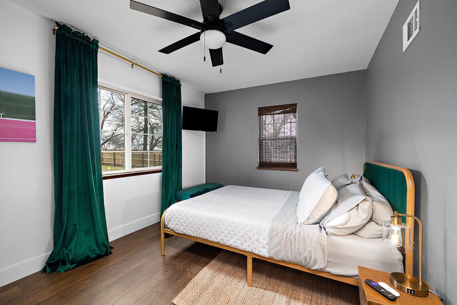 Un dormitorio con cama, ventilador de techo, televisión y ventana con cortinas.