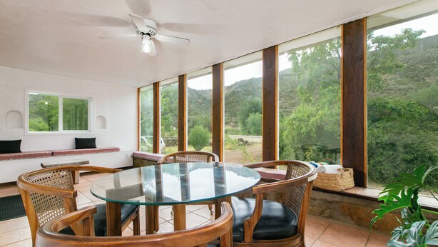 A dining area with a glass-top table and wooden chairs, large windows with a view of greenery, and a ceiling fan. - Relaxing Riverfront Taos Staycation, Pet Friendly! (Embudo)