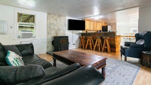 A living room with a sofa, a coffee table, and a TV mounted on the wall. There's a kitchen area with wooden cabinets and a bar stool. - Relaxing Riverfront Taos Staycation, Pet Friendly! (Embudo)
