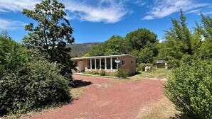 A single-story house with a red gravel driveway, surrounded by dense greenery and trees. - Relaxing Riverfront Taos Staycation, Pet Friendly! (Embudo)