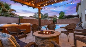 Une terrasse extérieure avec un foyer en pierre, des fauteuils en osier, un jacuzzi et des guirlandes lumineuses.