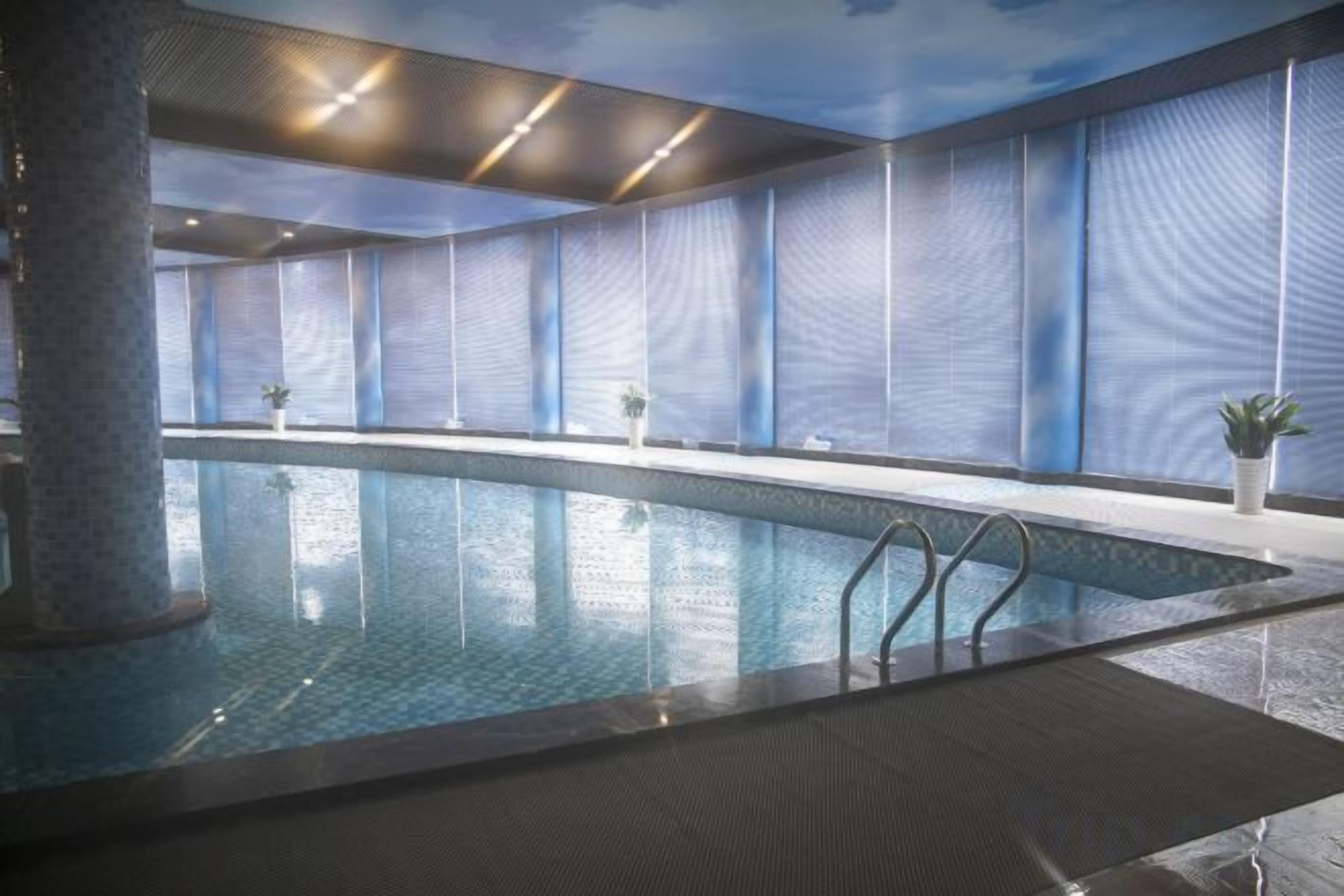 An indoor swimming pool with clear water, surrounded by walls with a wavy pattern, and a ceiling with recessed lighting.