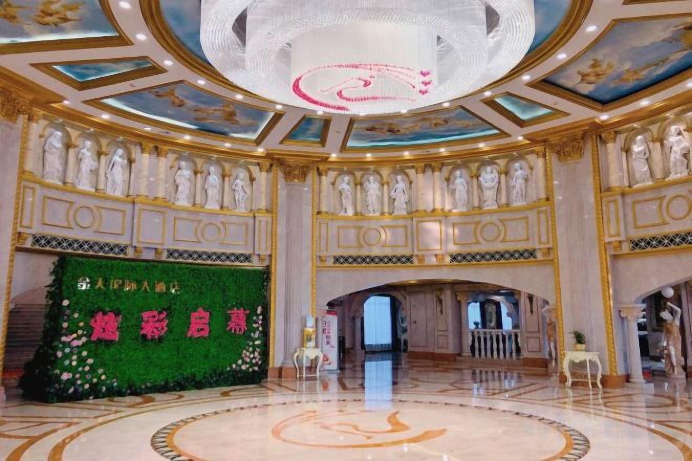 Um amplo saguão de hotel com piso de mármore circular, um lustre imponente e um painel floral verde com caracteres chineses.