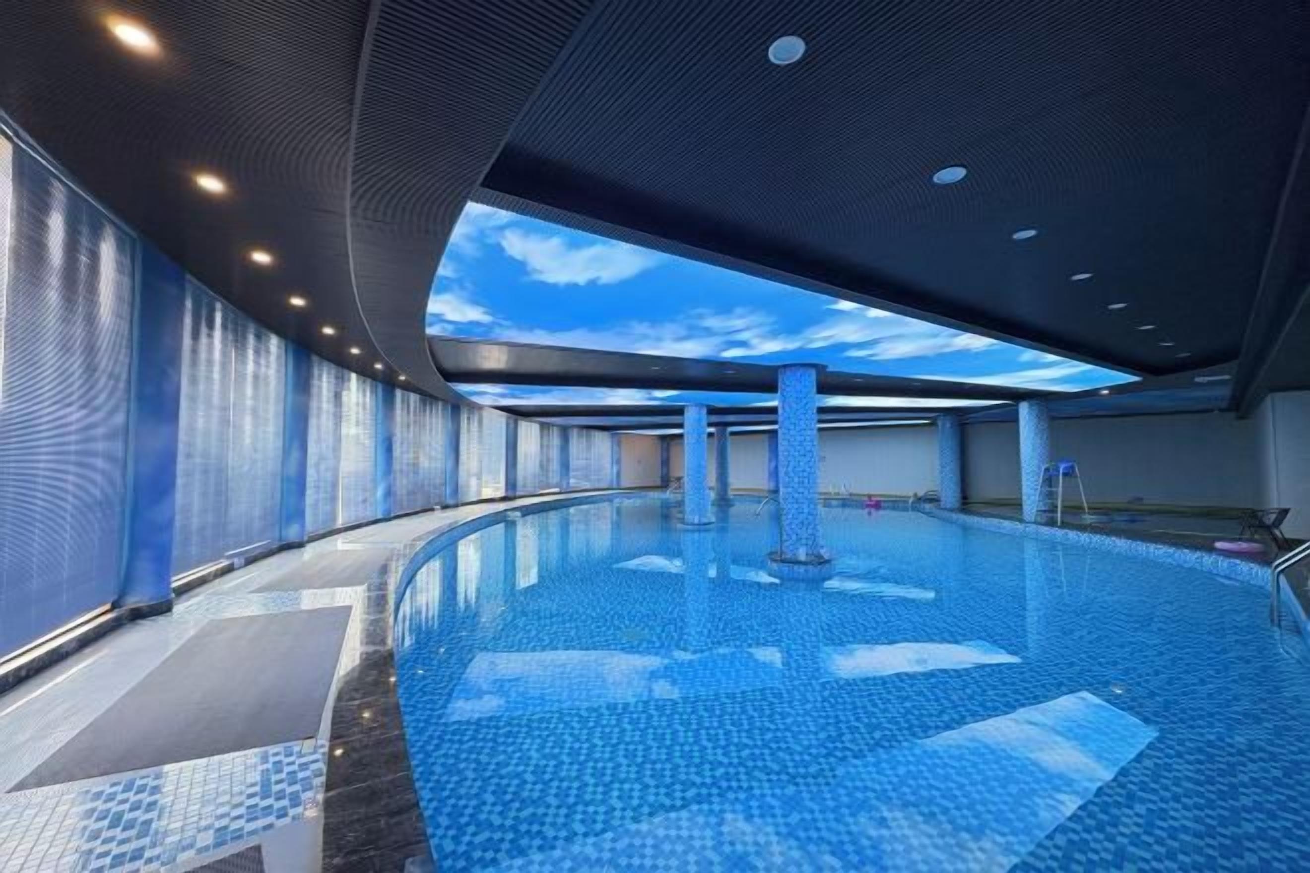 An indoor swimming pool with a curved design, a ceiling with a sky-like pattern, and a tiled floor.