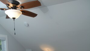 Ein Schlafzimmer mit einem Bett, einem Deckenventilator, einem Nachttisch mit Lampe, einer kleinen Pflanze und einer Wanddekoration.