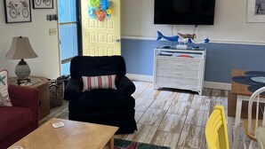 Een woonkamer met een zwarte fauteuil, een houten salontafel, een aan de muur bevestigde televisie en een deur met kleurrijke decoraties.