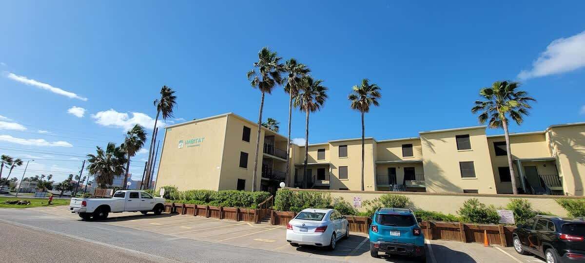 A multi-story hotel with a flat facade, palm trees, and a parking lot with several cars.