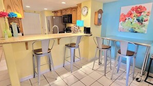 A kitchen with a bar area, equipped with a refrigerator, microwave, and sink. There are three bar stools and a clock on the wall.