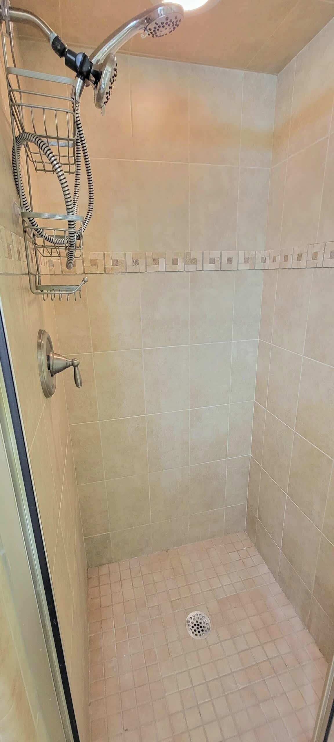 Une petite salle de douche carrelée, équipée d’un pommeau de douche à main, d’une étagère en métal et d’un drain.
