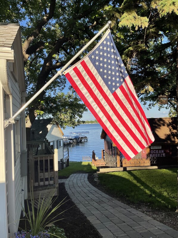 An American flag on a flagpole in front of a house with a dock and boats in the background.