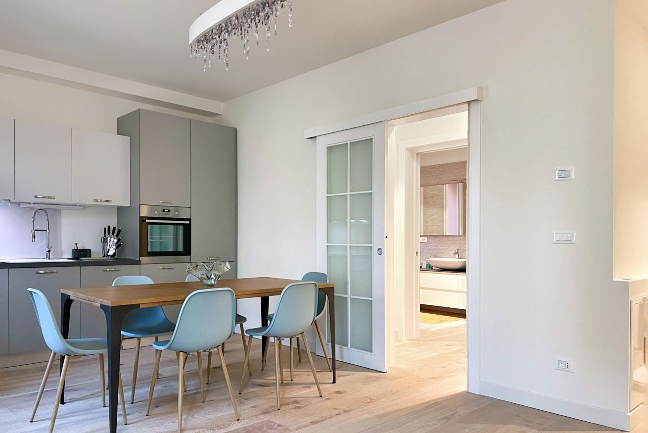 A modern kitchen with a dining area, featuring a wooden table and light blue chairs, a chandelier, and a sliding glass door leading to another room.